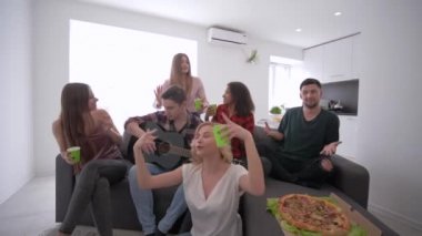 arkadaşlar eğlenceli dans ve gitar ile şarkı bir ev partisinde büyük bir modern dairede ellerinde plastik bardak ile kanepede oturan