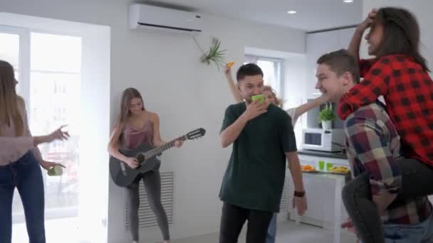 Веселые молодые друзья на домашней вечеринке веселятся и танцуют, пока девушка играет на гитаре в квартире — стоковое видео