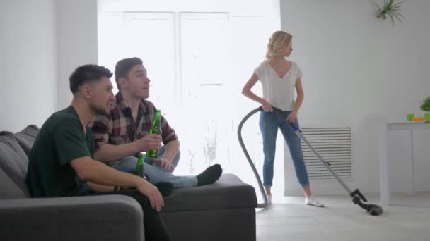 Amigos hombres viendo fútbol con cerveza en sus manos en el fondo de una chica nerviosa divertida con una aspiradora limpiando el apartamento — Vídeo de stock