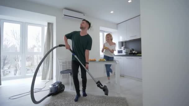 Jovens amantes engraçados está se divertindo na cozinha, homem feliz está dançando com aspirador de pó perto de mulher jovem que canta com uma concha como microfone — Vídeo de Stock