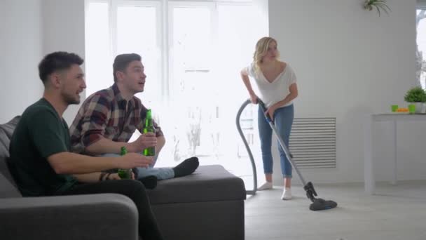 Male friends watching football with beer sitting on sofa on background of girl with a vacuum cleaner and then give each other a high five — Stock Video