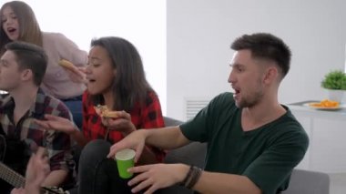 gülümseyen arkadaşlar pizza yemek ve ev partisinde kanepede oturan gitar ile şarkı lar