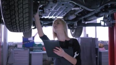 neşeli kadın servis istasyonunda bir hidrolik asansör üzerinde arabanın altında bir dijital tablet kullanarak teknik durumu kontrol eder