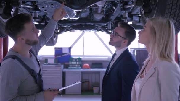 Auto mechanic adviseert jong paar klanten en houdt Klembord in handen staande onder de auto verhoogd op lift in auto service — Stockvideo
