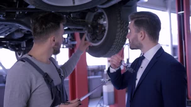 Mechaniker berät Kunden und macht sich Notizen in Klemmbrett, das unter Auto steht, das auf Aufzug im Autoservice angehoben wird — Stockvideo