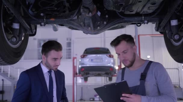 Mekaniker med servicebok inspektioner skick bil på hydraulisk lyft och prata med kund i bilservice — Stockvideo