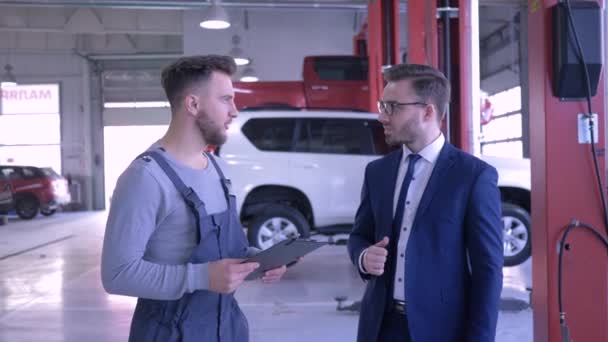 Entretien professionnel de voiture, mécanicien avec presse-papiers communique avec le client près de l'automobile sur ascenseur mécanique — Video