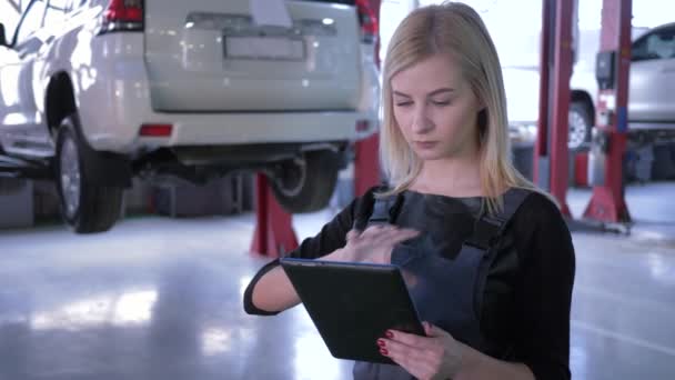 Профессиональная женщина-механик использует планшетный компьютер в автомастерской на фоне автомобилей на гидравлическом подъемнике — стоковое видео