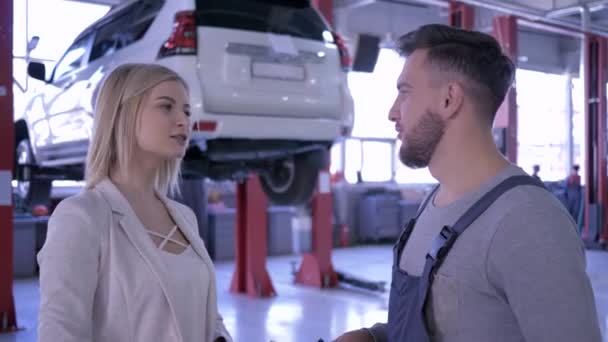 Professionelle Wartung, Automechaniker mit Schlüsseln, der Kundin die Hand schüttelt, blickt dann lächelnd in die Kamera vor dem Hintergrund des Autos auf dem Aufzug — Stockvideo