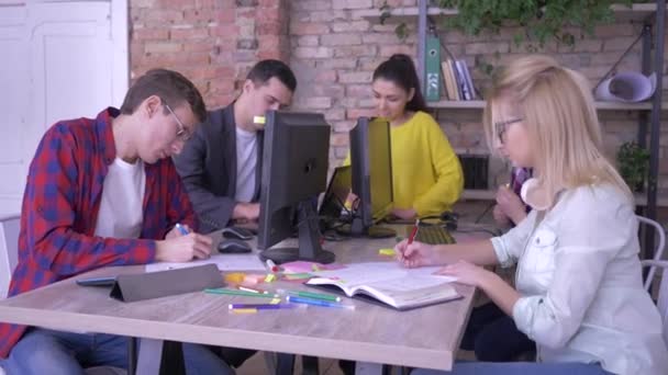 Aktivní mladí kanceláří pracovníci píší poznámky s nápady v poznámkových blocích a barevných nálepkami sedícího u stolu v moderní kanceláři — Stock video