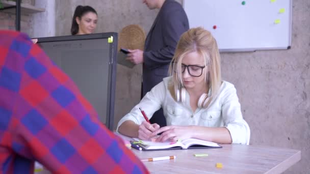 Empresaria con gafas sentada en el escritorio escribiendo notas en un cuaderno y pegamentos pegatinas de colores en la computadora en la oficina moderna — Vídeos de Stock