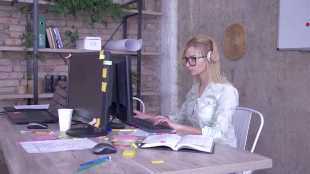 Arbeiterin mit Brille und Kopfhörer arbeitet am PC in einem modernen kreativen Büro — Stockvideo