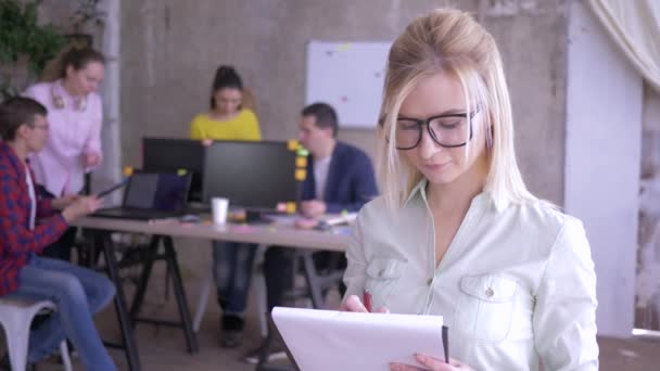 Gelukkige vrouwelijke kantoormedewerker in glazen maken van notities op papier en dan glimlachend op de camera — Stockvideo