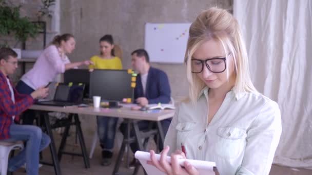 Porträt einer glücklichen Büroangestellten mit Brille, die Notizen auf Papier macht und dann in die Kamera lächelt — Stockvideo