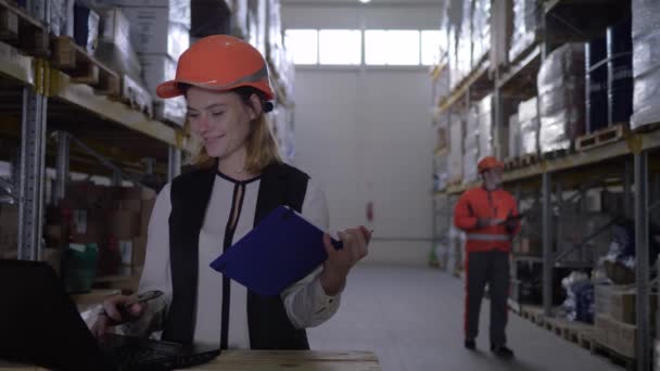 Zaměstnankyně v tvrdé čepici používat přenosný počítač a vytváří poznámky v poznámkovém bloku na pozadí pracovníka ze skladu — Stock video