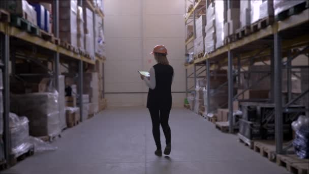 travailleuse industrielle en casque dur utilise tablette numérique tout en marchant entre les rangées de supports de stockage sur l'entrepôt d'usine 