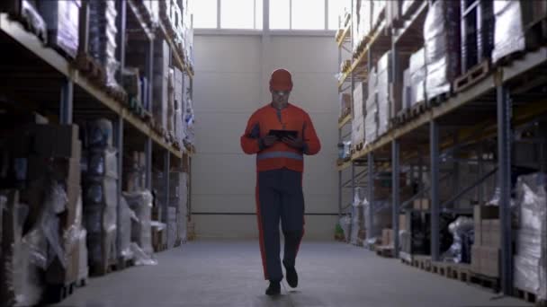 Worker wearing hard hat with tablet computer in warehouse walks through rows of storage racks with merchandise — Stock Video