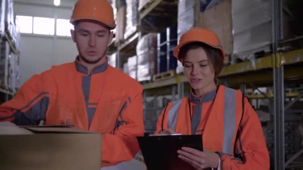 Empleados de almacén en ropa de trabajo y cascos discutir el trabajo y toma notas cerca de cajas — Vídeos de Stock