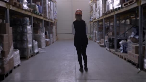 Woman worker in hard hat uses digital tablet on factory warehouse between rows of storage racks with merchandise — Stock Video