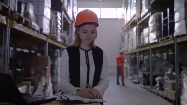 travailleur femme en casque dur utiliser ordinateur portable et prend des notes dans le bloc-notes dans l'entrepôt 