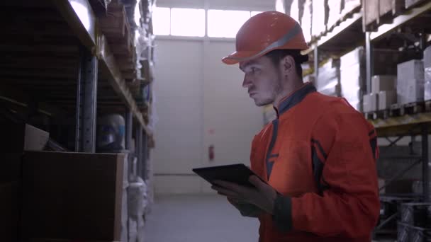 Νέος εργαζόμενος αποθήκη σε πορτοκαλί σκληρό καπέλο κοντά σε μεγάλα ράφια με αγαθά χρησιμοποιεί υπολογιστή tablet — Αρχείο Βίντεο