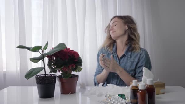 Jovem espirra, uma menina adulta é alérgica a flores enquanto se senta à mesa com medicamentos e limpa o nariz com guardanapo — Vídeo de Stock