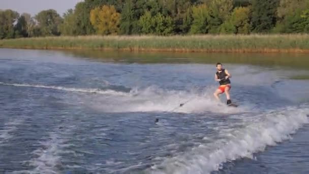 Aktív életmód, wakeboarder ember lovagol a fedélzeten mögött motorcsónak a folyón vízzel fröccsenő háttérben a nád-és fák — Stock videók