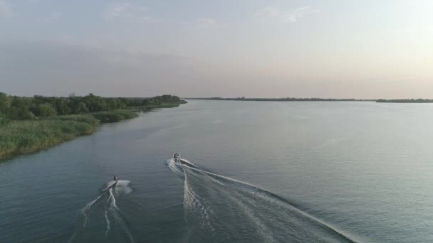 Vista aérea deportista sostiene asa de cuerda y paseos en su wakeboard a lo largo del río detrás de un barco a motor al aire libre en cámara lenta — Vídeos de Stock