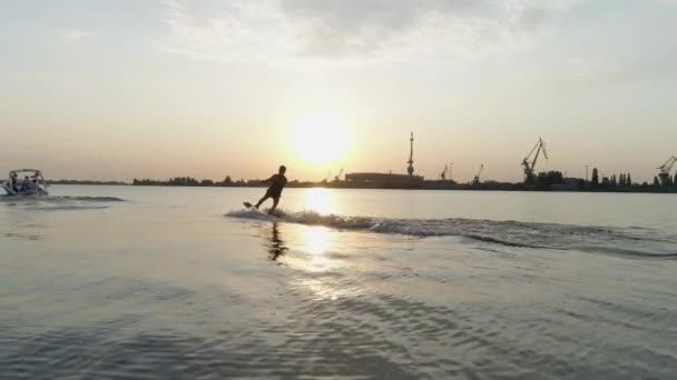 Vista aérea, surfista hombre paseos en wakeboard a lo largo del río detrás de la lancha a motor y sostiene la manija de la cuerda en las manos durante la puesta del sol — Vídeos de Stock