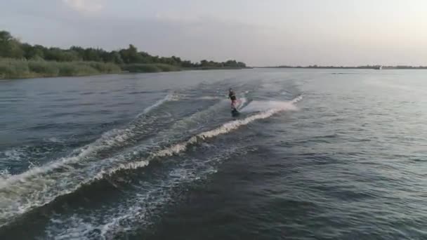 Безпілотний постріл, спортсмен їде на борту позаду моторного човна з бризками води вздовж річки під час літніх канікул на природі — стокове відео