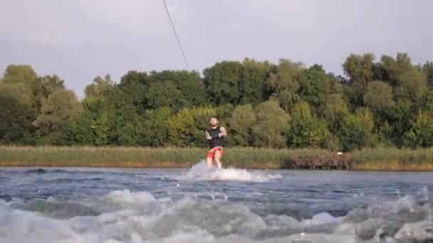 Happy wakeboarder rides on board behind motorboat with splashes and holds rope handle, sporting man training on river during weekend — Stock Video