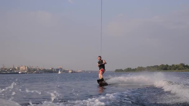 활짝 웃는 서퍼 남자는 슬로우 모션에 포트와 푸른 하늘의 배경에 물이 야외에서 튀는 모터 보트 뒤에 보드에 타고 — 비디오