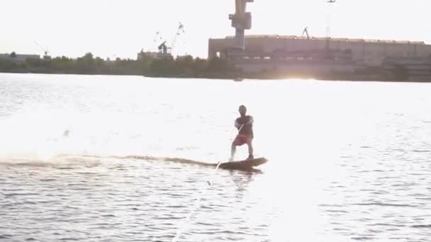 Esportes pessoa na luz do sol passeios no wakeboard atrás de lancha com salpicos de água ao longo do rio no fundo da natureza em câmera lenta — Vídeo de Stock