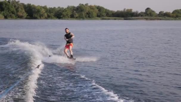 Water extreme sport, wakeboarder man holds rope handle and rides on board on river in slow motion with splashes on background of nature — Stock Video