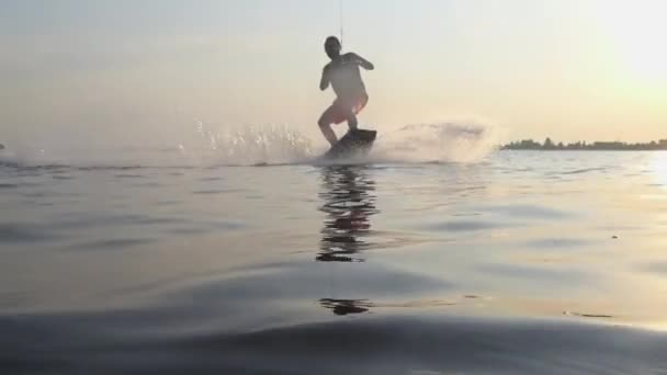 Water sport, wakeboarder man rides on board behind motorboat on river and make splashes on camera lens on background of golden sun and blue sky — Stock Video