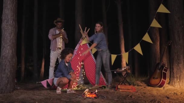 Кемпинг, красивая жена и ее веселый муж поставить палатку после отдыха в лесу в то время как маленькая дочь жарит зефир у костра — стоковое видео