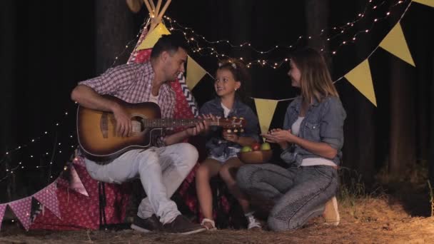 Familia en el bosque, los padres jóvenes con el niño hembra se divierten tocando la guitarra y comiendo frutas en el picnic nocturno en el bosque telón de fondo de wigwam — Vídeo de stock