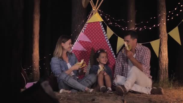 Οικογενειακό πικ-νικ, γονείς και γοητευτική κόρη διασκεδάζουν τρώγοντας φρούτα ενώ έχουν σνακ στο δείπνο στο δάσος στο φόντο της καλυβοσκηνή — Αρχείο Βίντεο