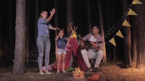 Glücklicher Vater spielt Gitarre für seine Familie, Mutter und Tochter haben Spaß am Lagerfeuer am Wochenende im Wald vor dem Hintergrund von Wigwam — Stockvideo