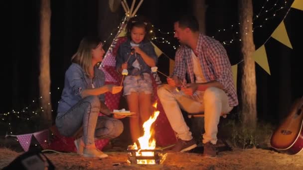 Marito e moglie con bambino trascorrono felicemente del tempo in un'atmosfera familiare mangiando marshmallow fritti al falò durante il picnic nella foresta notturna vicino a wigwam — Video Stock