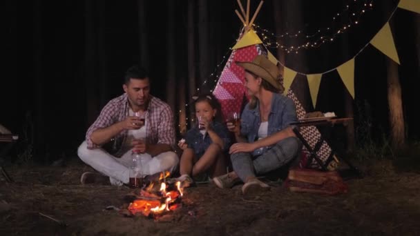 Loving relatives, mom with dad and little daughter make a toast with glasses of juice in woods around campfire during night picnic on wigwam background — ストック動画