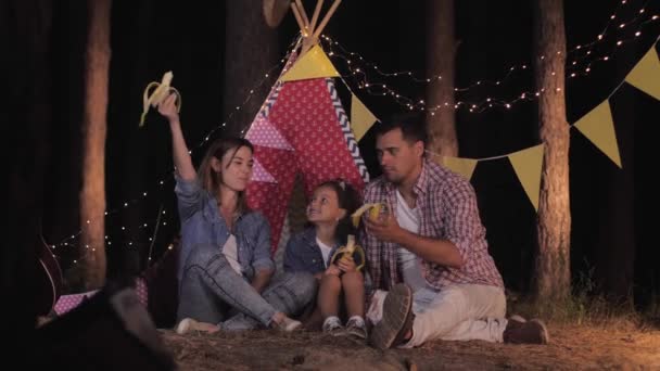 Pasatiempo de los padres y el niño, hermosa niña descansando en el picnic con sus padres cariñosos comer refrescos para el fin de semana familiar en el bosque en el fondo de wigwam — Vídeos de Stock