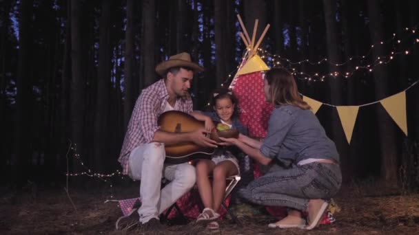 若い母親は家族のための果物とプレートを持って来る、両親と楽しい子供の女の子は、夕方のキャンプ旅行中にギターでウィグワムの近くで時間を過ごす — ストック動画