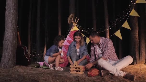 Пикник ночью, веселая семья мама папа и дочь разжигают костёр в лесу на отдыхе за городом на фоне wigwam — стоковое видео