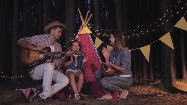 Tijd samen, vrolijke vader speelt gitaar en zingt door vuur op picknick in het bos voor vrolijke vrouw en mooie dochter, familie klappen handen op de achtergrond wigwam — Stockvideo