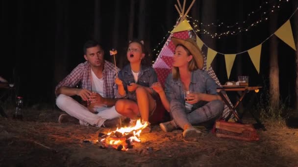 Encantadora filha e pais alegres conversando em torno de fogo beber suco e fritar marshmallows no piquenique na floresta noturna durante as férias de verão — Vídeo de Stock