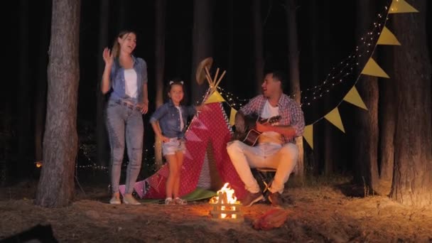 Семейное веселье, счастливый отец играет на гитаре для мамы с дочкой, которые танцуют у костра в летние выходные в лесу на фоне вигвамов — стоковое видео