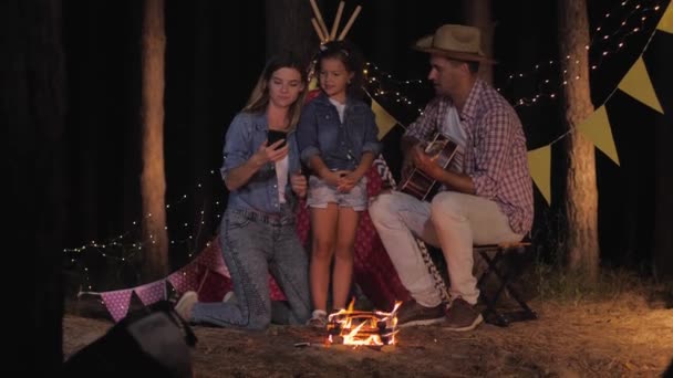 Family selfie, smiling mom and dad with their charming daughter take photos in woods around campfire during nights rest — Stock Video