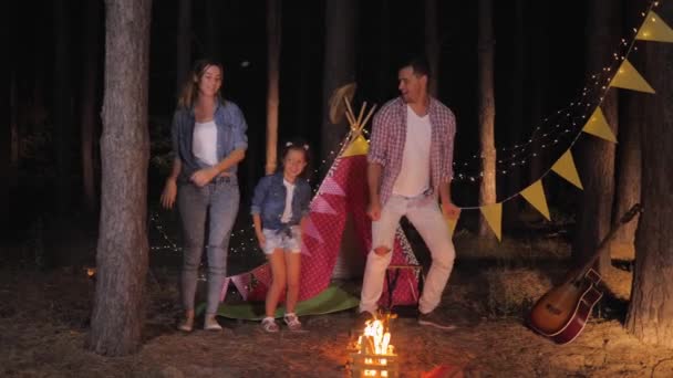 Семейный отдых, счастливые родители с радостной дочерью весело танцуют вместе на пикнике в лесу во время кемпинга в летний фон wigwam и костер — стоковое видео