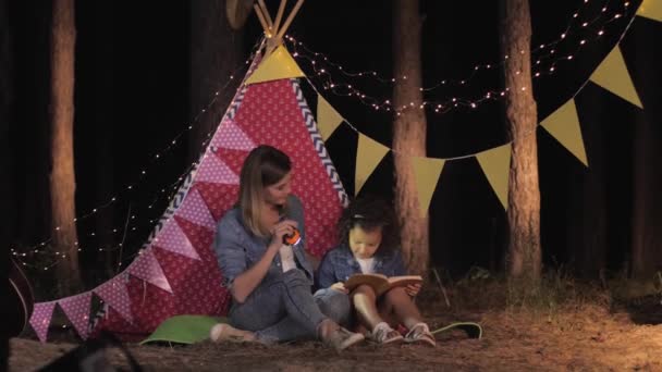 Familia feliz, madre cariñosa y hermosa hija leer historias interesantes en el libro en el bosque durante el fin de semana al aire libre — Vídeo de stock